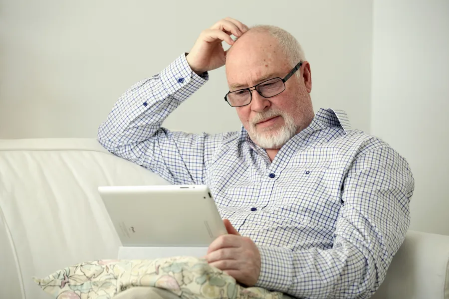 En mann som sitter i en sofa, ser på et nettbrett og klør seg i hodet.