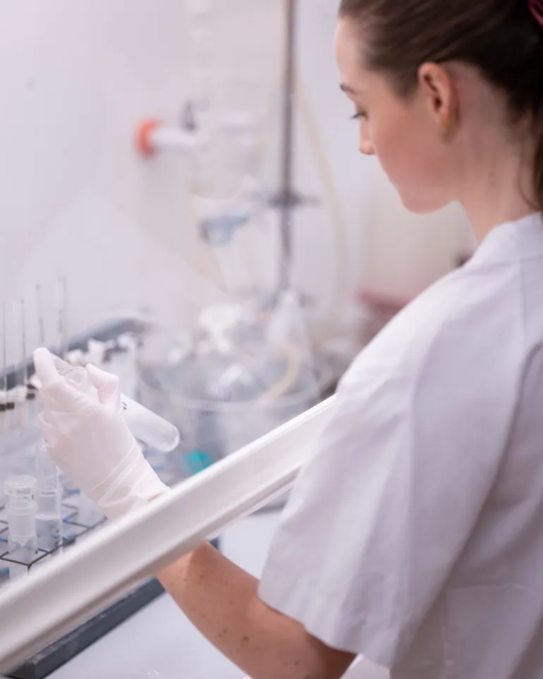 Laboratoriearbeider i sterilbenk holder et prøverør med klar væske, omgitt av annet analyseutstyr.