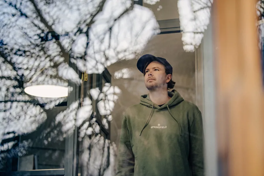 Mann står innendørs og ser ut av et vindu med et tankefullt ansiktsuttrykk.