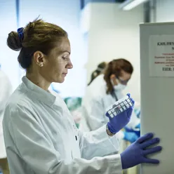 Mennesker som jobber i et laboratorium. De har på frakker og hansker, og hun ene titter på prøver i et fryseskap.