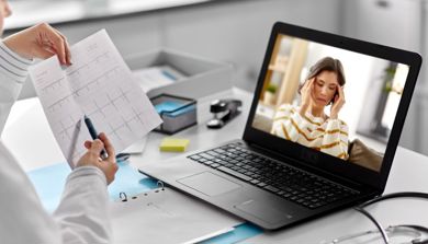 En laptop som viser et bilde av en dame som holder seg til tinningene. Foran skjermen er to hender som holder i et ark.