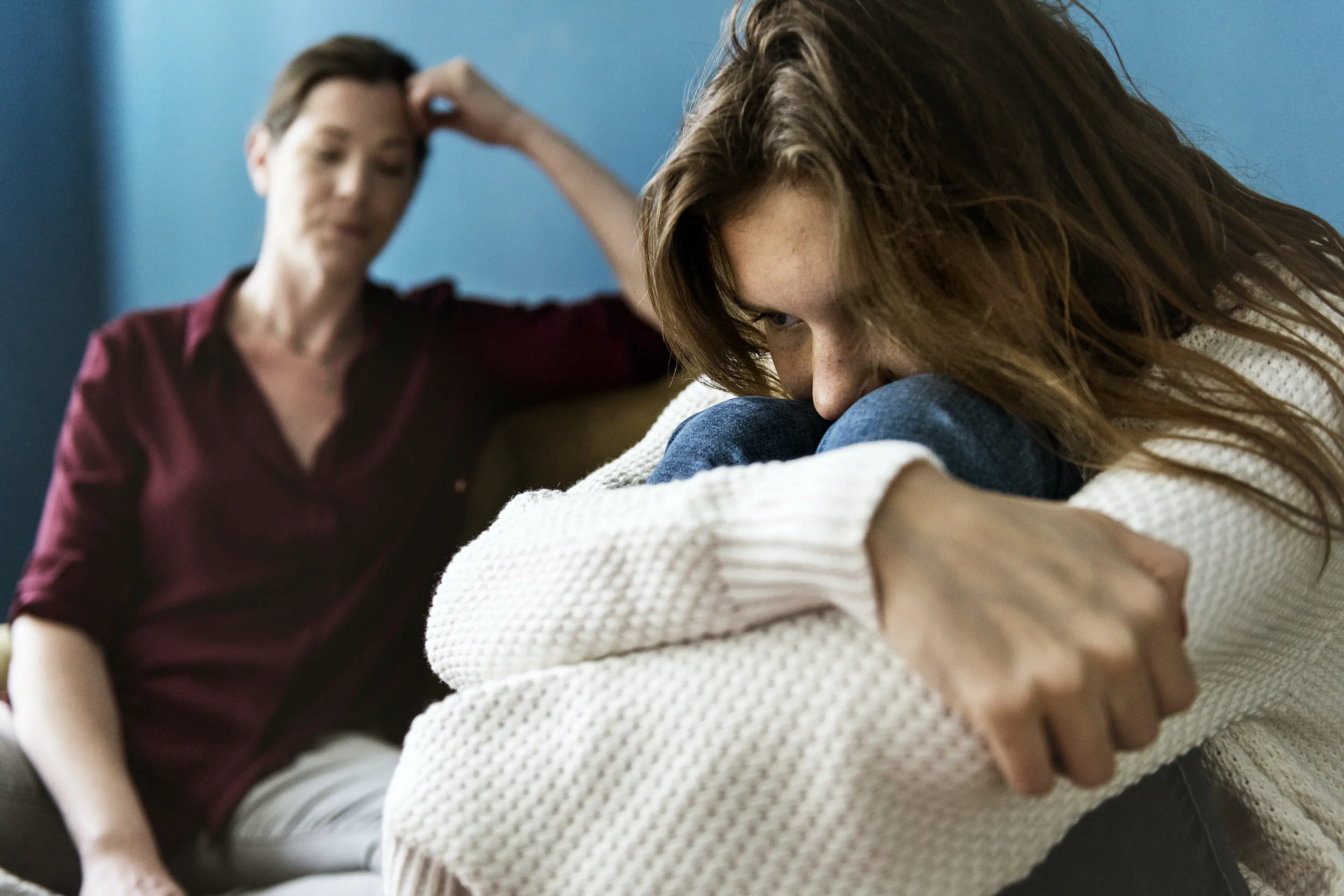 Trist jente som sitter i fosterstilling på en sofa med moren sin