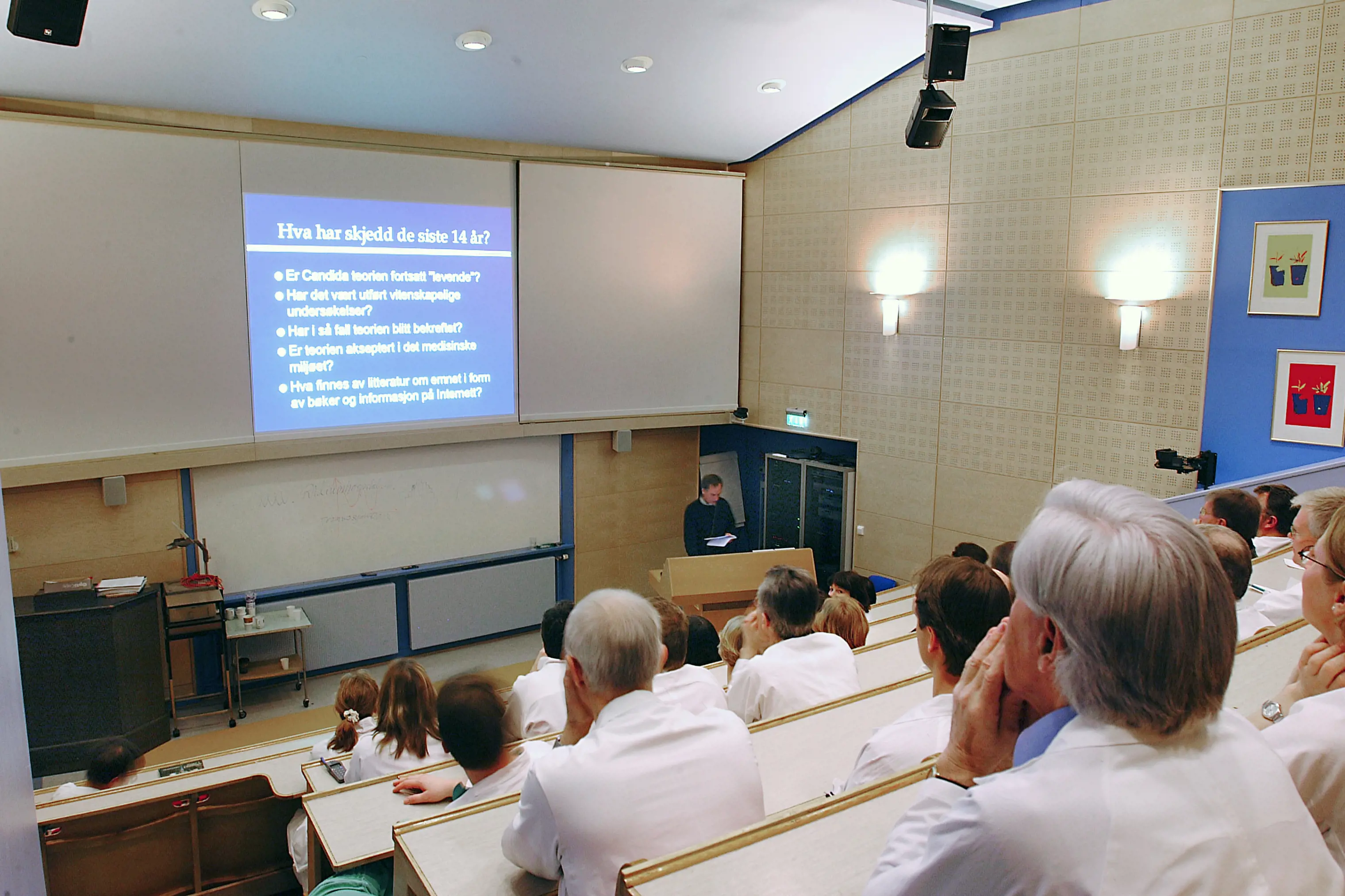 En gruppe mennesker i hvite frakker følger en forelesning i et  auditorium