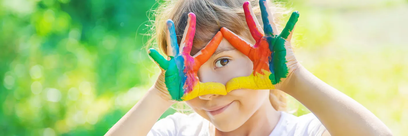 Et barn med maling på fingrene, som holder hendene som et kikkhull mot øyet