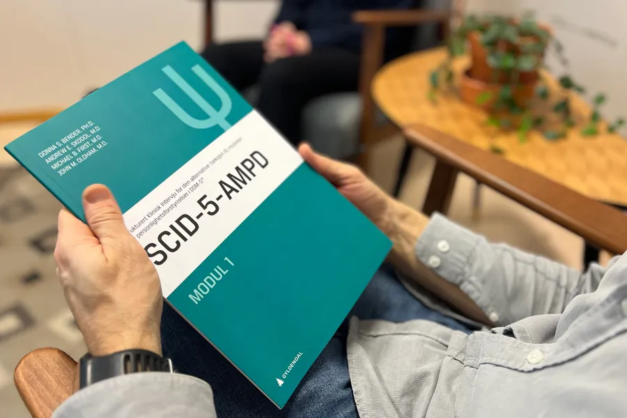 Utredningsverktøyet SCID-5-AMPD holdes en av terapeut i terapirommet, i bakgrunnen sitter en pasient.