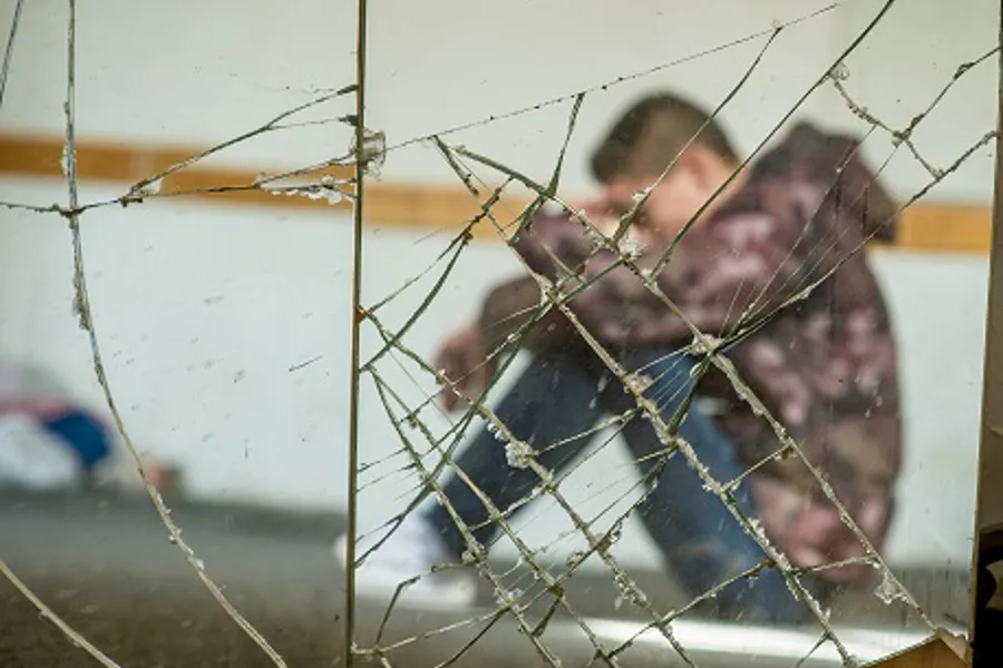 En ung gutt med hettejakke sitter med hodet bøyd bak knust glass