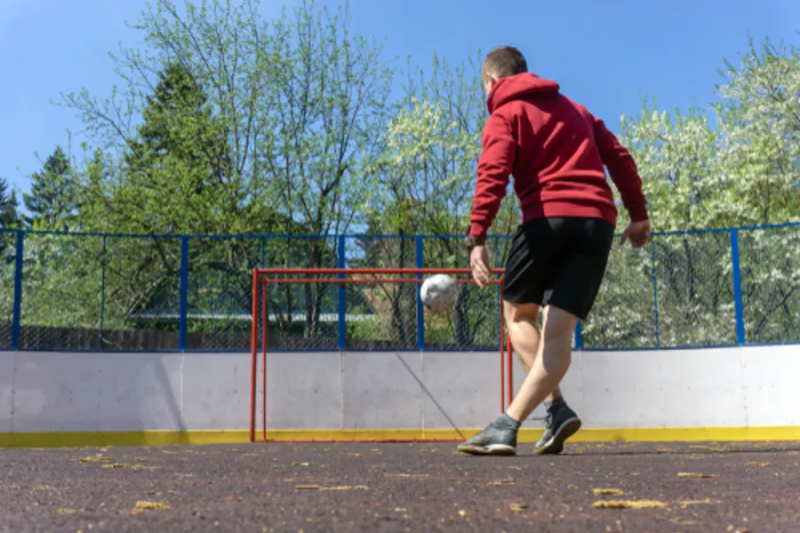 Ungdomsgutt med sort shorts og rød hettegenser sparker ball mot et fotballmål.